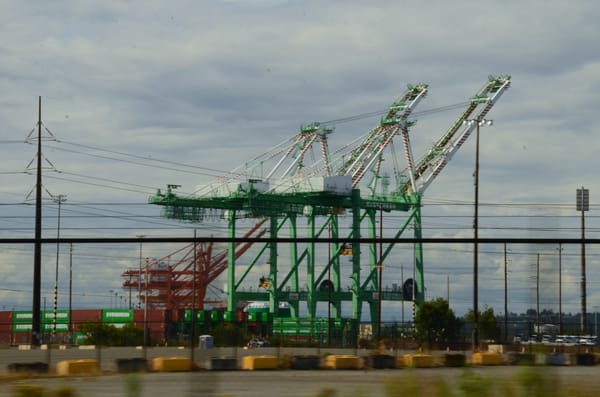 Two green cargo cranes in the distance with a freight train and cargo containers arranged behind a chain link fence. (Photo taken by Chris Karnes)